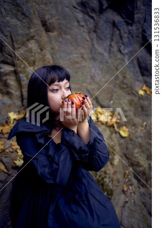 woman in a black long dress sits on black rocks on Halloween holding a pumpkin-shaped cup, a modern witch 131536833