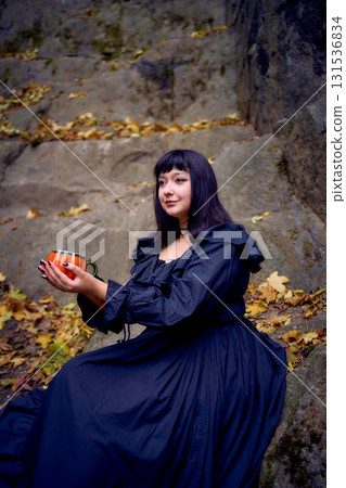 woman in a black long dress sits on black rocks on Halloween holding a pumpkin-shaped cup, a modern witch 131536834