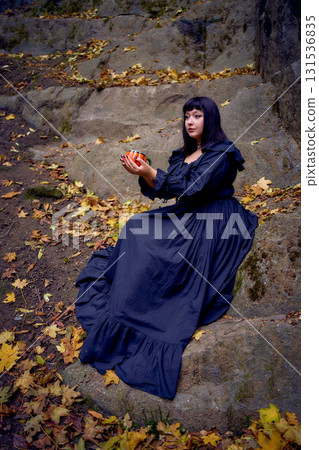 woman in a black long dress sits on black rocks on Halloween holding a pumpkin-shaped cup, a modern witch woman in a black long dress sits on black rocks on Halloween holding a pumpkin-shaped cup, a modern witch 131536835