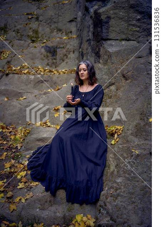 woman in a black long dress sits on black rocks on Halloween holding a burning candle, performs a witch ritual, modern witch woman in a black long dress sits on black rocks on Halloween holding a burning candle, performs a witch ritual, modern witch 131536836