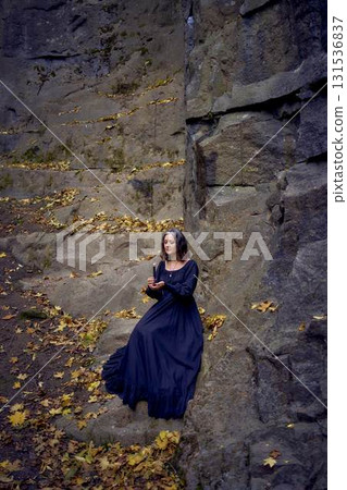 woman in a black long dress sits on black rocks on Halloween holding a burning candle, performs a witch ritual, modern witch woman in a black long dress sits on black rocks on Halloween holding a burning candle, performs a witch ritual, modern witch 131536837