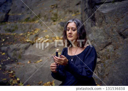 woman in a black long dress sits on black rocks on Halloween holding a burning candle, performs a witch ritual, modern witch woman in a black long dress sits on black rocks on Halloween holding a burning candle, performs a witch ritual, modern witch 131536838