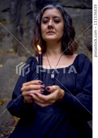 woman in a black long dress sits on black rocks on Halloween holding a burning candle, performs a witch ritual, modern witch woman in a black long dress sits on black rocks on Halloween holding a burning candle, performs a witch ritual, modern witch 131536839