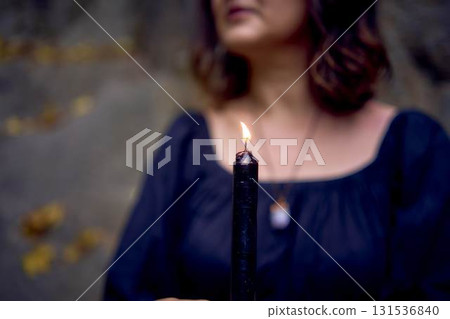 woman in a black long dress sits on black rocks on Halloween holding a burning candle, performs a witch ritual, modern witch 131536840