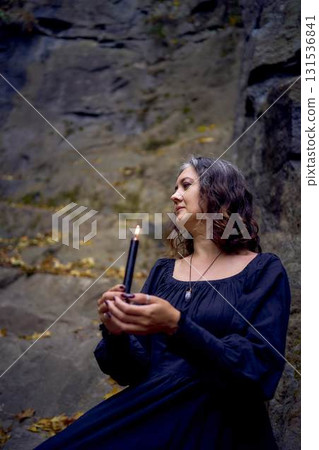 woman in a black long dress sits on black rocks on Halloween holding a burning candle, performs a witch ritual, modern witch 131536841