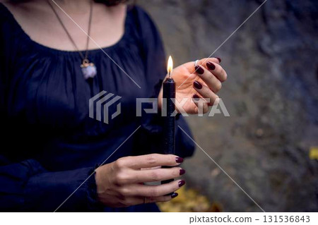 woman in a black long dress sits on black rocks on Halloween holding a burning candle, performs a witch ritual, modern witch woman in a black long dress sits on black rocks on Halloween holding a burning candle, performs a witch ritual, modern witch 131536843