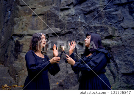 two women in the black long dresses near black rocks on Halloween holding a burning candle, performing a witch ritual, modern witch 131536847
