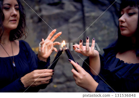 two women in the black long dresses near black rocks on Halloween holding a burning candle, performing a witch ritual, modern witch two women in the black long dresses near black rocks on Halloween holding a burning candle, performing a witch ritual, modern witch 131536848