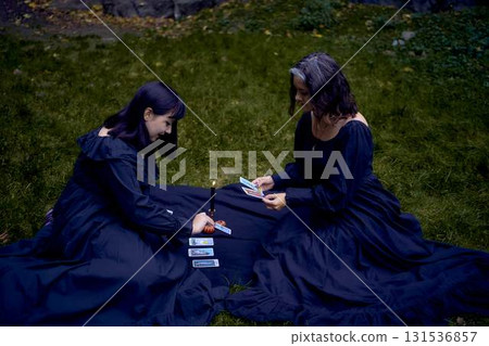 two women in black long dresses sitting on the black rocks on Halloween, reading tarot cards, performing a witch ritual, modern witch two women in black long dresses sitting on the black rocks on Halloween, reading tarot cards, performing a witch ritual, modern witch 131536857