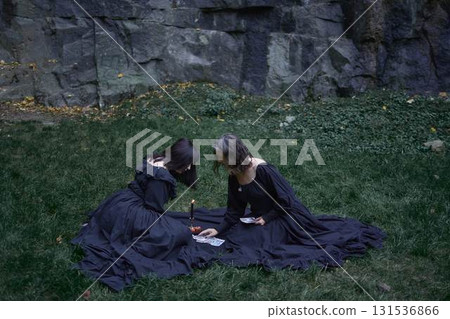 two women in black long dresses sitting on the black rocks on Halloween, reading tarot cards, performing a witch ritual, modern witch two women in black long dresses sitting on the black rocks on Halloween, reading tarot cards, performing a witch ritual, modern witch 131536866