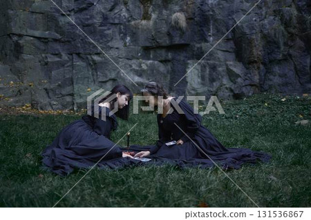 two women in black long dresses sitting on the black rocks on Halloween, reading tarot cards, performing a witch ritual, modern witch 131536867