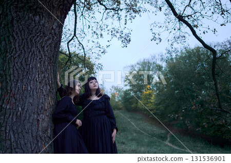 Two women in the long black dresses perform a historical reenactment of the Edwardian era dancing lovers on a hill near a majestic oak tree for Halloween. 131536901