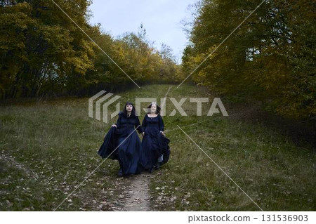 Two women in the long black dresses perform a historical reenactment of the Edwardian era dancing lovers on a hill near a majestic oak tree for Halloween. 131536903