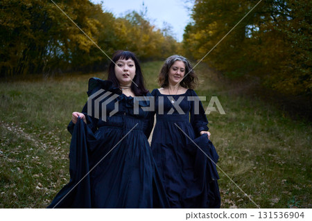 Two women in the long black dresses perform a historical reenactment of the Edwardian era dancing lovers on a hill near a majestic oak tree for Halloween. Two women in the long black dresses perform a historical reenactment of the Edwardian era dancing lovers on a hill near a majestic oak tree for Halloween. 131536904