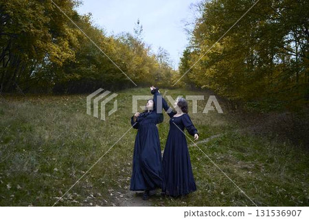Two women in the long black dresses perform a historical reenactment of the Edwardian era dancing lovers on a hill near a majestic oak tree for Halloween. 131536907