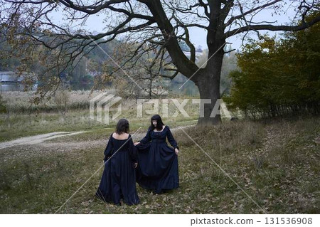Two women in the long black dresses perform a historical reenactment of the Edwardian era dancing lovers on a hill near a majestic oak tree for Halloween. 131536908