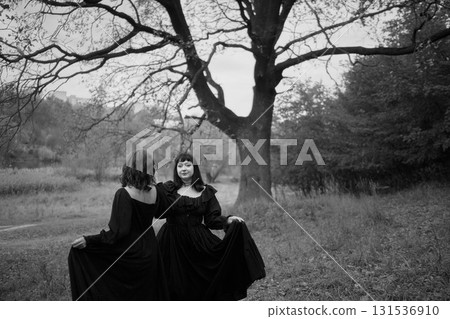 Two women in the long black dresses perform a historical reenactment of the Edwardian era dancing lovers on a hill near a majestic oak tree for Halloween. 131536910