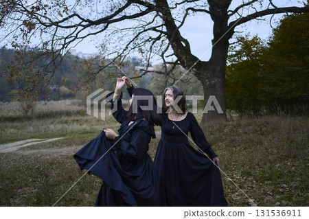 Two women in the long black dresses perform a historical reenactment of the Edwardian era dancing lovers on a hill near a majestic oak tree for Halloween. Two women in the long black dresses perform a historical reenactment of the Edwardian era dancing lovers on a hill near a majestic oak tree for Halloween. 131536911