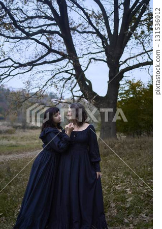 Two women in the long black dresses perform a historical reenactment of the Edwardian era dancing lovers on a hill near a majestic oak tree for Halloween. 131536912
