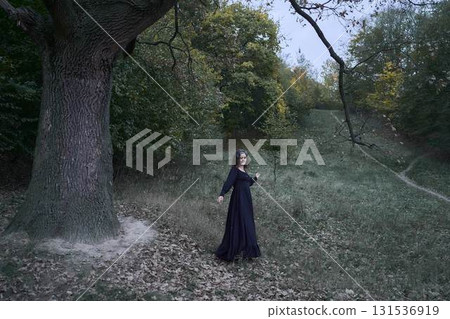 a women in the long black dresses perform a historical reenactment of the Edwardian era on a hill near a majestic oak tree for Halloween. 131536919