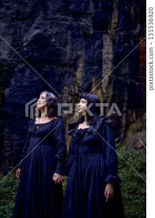 two women in the black long dresses sitting on black rocks on Halloween, performing a witch ritual, modern witch two women in the black long dresses sitting on black rocks on Halloween, performing a witch ritual, modern witch 131536920