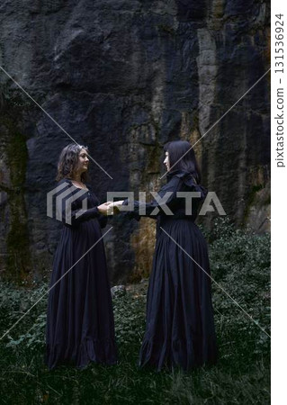 two women in the black long dresses sitting on black rocks on Halloween, performing a witch ritual, modern witch two women in the black long dresses sitting on black rocks on Halloween, performing a witch ritual, modern witch 131536924