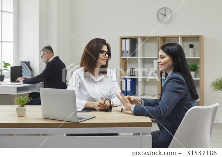 Portrait of attractive, young women sitting at desk against the background of their colleague, in spacious, modern office. Discussion of working moments. Portrait of attractive, young women sitting at desk against the background of their colleague, in spacious, modern office. Discussion of working moments. 131537186