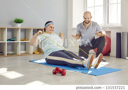 Friendly nurse helping fat woman doing stretching exercises with rubber band sitting on the floor 131537221