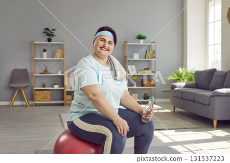 Fat woman wearing sportswear sitting on fit ball with a bottle of water after fit exercises at home Fat woman wearing sportswear sitting on fit ball with a bottle of water after fit exercises at home 131537223