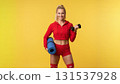 fitness woman stands confidently with a dumbbell in one hand and a yoga mat in the other. She wears a red workout outfit and smiles warmly radiating energy and positivity. 131537928