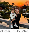 asian woman stands gracefully by a serene pond holding a young boy close as the sun sets behind a traditional garden setting. The colors of dusk reflect on the water. 131537940