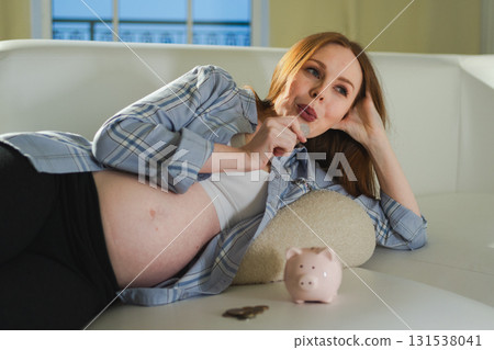 Pregnancy motherhood saving money for future investment. Pregnant woman with big belly putting coin into piggy bank at home. Pregnancy girl with piggy bank. Maternity benefit family planning concept 131538041