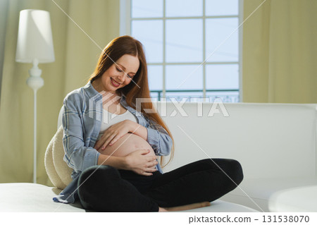 Pregnancy motherhood people expectation future. Pregnant woman touching big belly sitting on couch at home. Girl hugging her tummy enjoying pregnancy. Maternity tenderness parenthood new life concept Pregnancy motherhood people expectation future. Pregnant woman touching big belly sitting on couch at home. Girl hugging her tummy enjoying pregnancy. Maternity tenderness parenthood new life concept 131538070