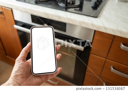 Hand holding smartphone with white blank screen near modern kitchen oven. Woman operating through mobile application. Smart home. Close up Hand holding smartphone with white blank screen near modern kitchen oven. Woman operating through mobile application. Smart home. Close up 131538087