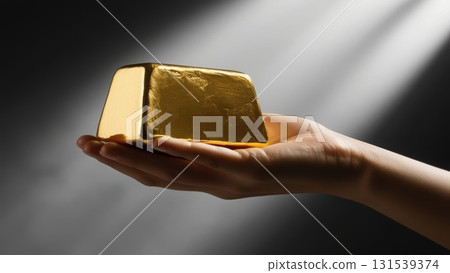 Hand holding a heavy solid gold ingot against a dark blurred background emphasizing richness 131539374