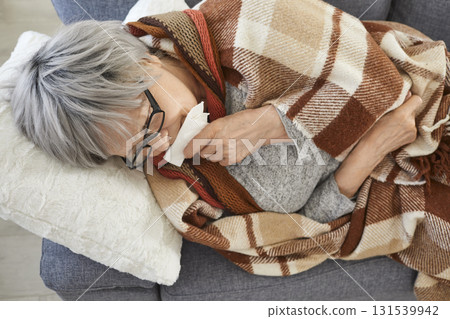 Senior sick woman lying on sofa at home wrapped in a blanket suffering from runny nose. Senior sick woman lying on sofa at home wrapped in a blanket suffering from runny nose. 131539942