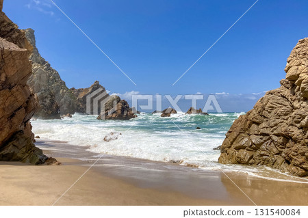 Ursa beach near Cabo da Roca features rugged cliffs, golden sand, and crashing waves under a clear blue sky 131540084