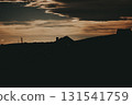 Silhouette of houses on a hill against a dramatic sunset sky. Silhouetted house roof perched on a hill, with a stunning sunset sky as the backdrop, creating a serene and picturesque scene. Rural land. 131541759