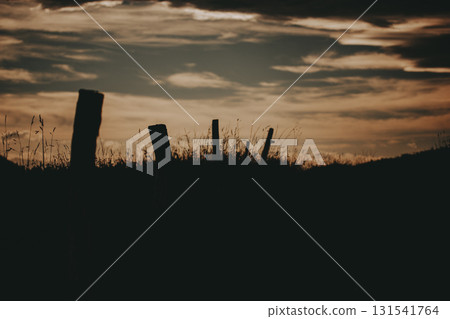 Silhouette of fence posts against a dramatic sunset sky. A moody serene landscape captures the silhouette of weathered fence posts set against a vibrant, cloud-filled sunset, creating a tranquil scene Silhouette of fence posts against a dramatic sunset sky. A moody serene landscape captures the silhouette of weathered fence posts set against a vibrant, cloud-filled sunset, creating a tranquil scene 131541764