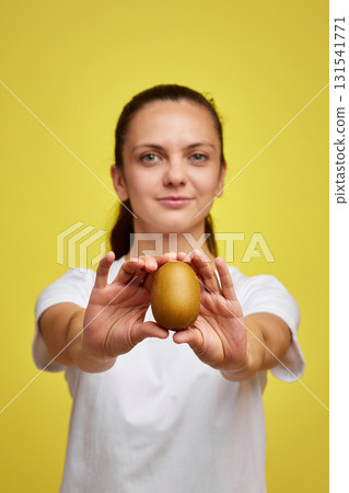 Happy woman in white t-shirt holding kiwi on studio yellow background, focus on kiwi Happy woman in white t-shirt holding kiwi on studio yellow background, focus on kiwi 131541771