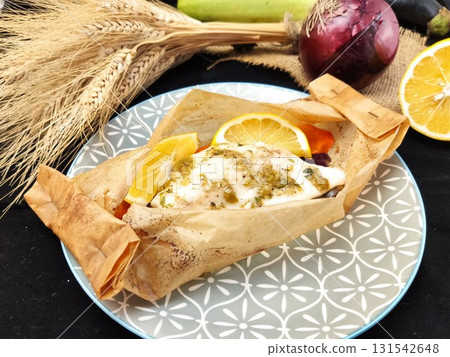 Baked chicken breast in a parcel with lemon wedges on top. The dish sits on a gray patterned plate. In the background are ears of wheat, red onion, and vegetables. 131542648
