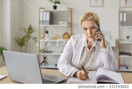 Focused mature woman working at desk with laptop, taking notes while making work calls on phone. Focused mature woman working at desk with laptop, taking notes while making work calls on phone. 131543602