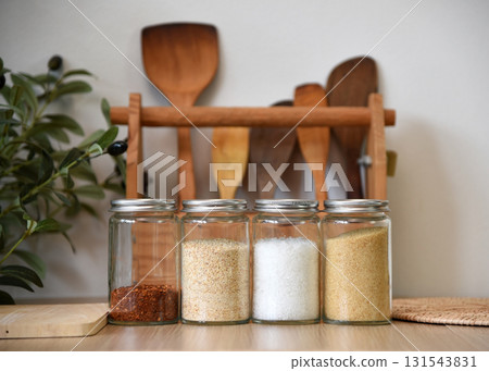 A clear jar that holds seasonings for cooking inside. On the wooden table in the kitchen There are cooking equipment in the background. 131543831