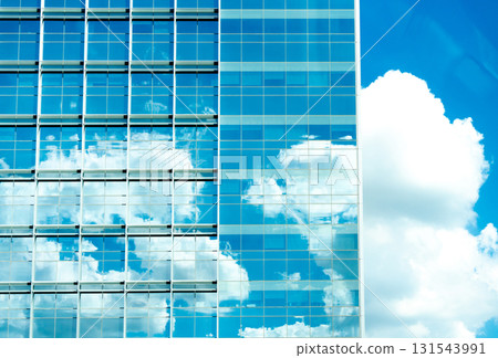 Glass office facade reflects blue sky and cumulus clouds, merging city and nature. 131543991