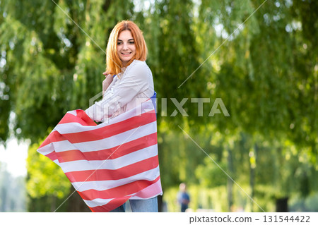Happy young woman posing with USA national flag standing outdoors in summer park. Positive girl with United States banner outdoors. Happy young woman posing with USA national flag standing outdoors in summer park. Positive girl with United States banner outdoors. 131544422