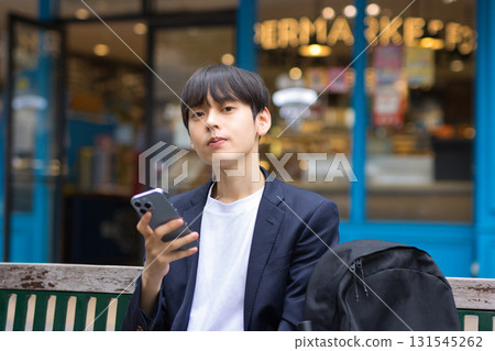 A male businessman in his twenties sitting on a bench and looking at his smartphone A male businessman in his twenties sitting on a bench and looking at his smartphone 131545262