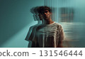 Studio shot of a young man wearing a white t shirt standing behind textured glass, creating wavy light patterns and shadows on his body and the background, giving a modern and artistic feel 131546444