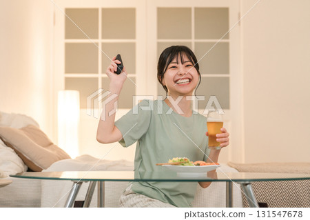 Young Asian woman laughing while watching TV and having a drink at dinner Young Asian woman laughing while watching TV and having a drink at dinner 131547678