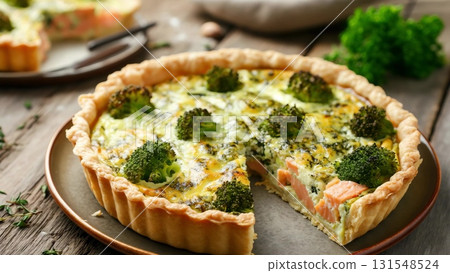 Golden brown quiche featuring salmon and broccoli, sliced for revealing creamy filling and delicate pastry layers atop rustic wooden surface 131548524