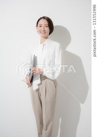 A beautiful woman in office casual wear and a blouse standing with a laptop on a white background, looking at the camera A beautiful woman in office casual wear and a blouse standing with a laptop on a white background, looking at the camera 131552996